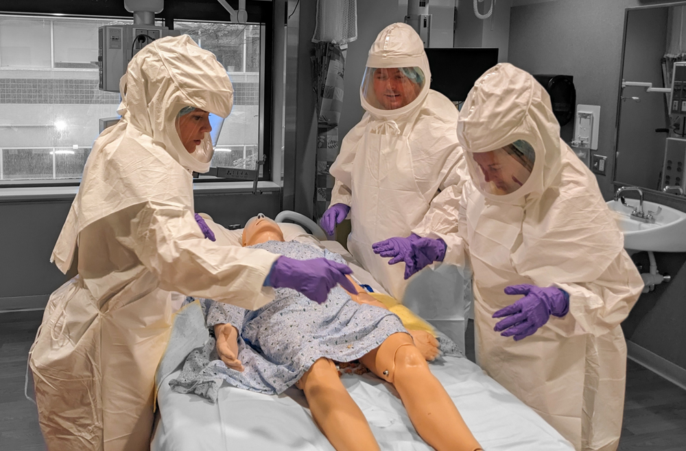 Three staff members in full-body personal protective equipment surround a dummy on a bed in a hospital room.